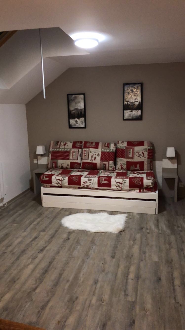 A cozy sitting area features a fold-out couch adorned with a red and white patterned cover. Two framed artworks are hung on the wall above the couch. A small, fluffy rug is placed on the wooden floor, enhancing the space's comfort.