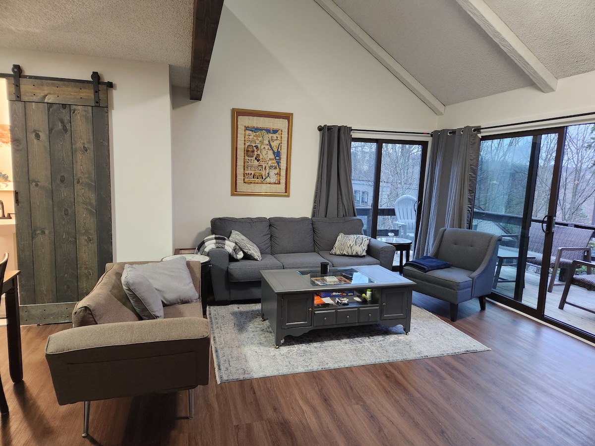 A living area features a mix of comfortable seating arrangements, including a gray sectional sofa and a stylish armchair. A coffee table is centrally located, set atop a light area rug. Large glass doors lead to a deck area, framed by two windows offering views of the outdoors.