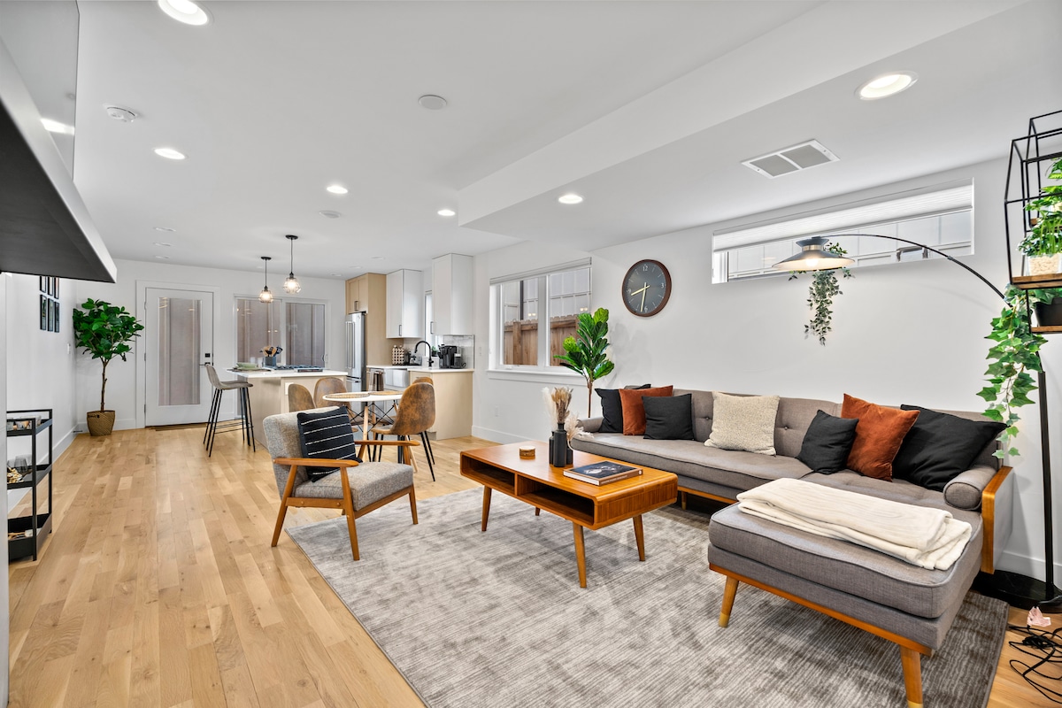 A modern open-concept living area features a comfortable sectional sofa accented with orange and brown cushions. A wooden coffee table sits atop a soft area rug. Natural light enters through large windows, illuminating the space where a dining table and chairs are visible in the background.