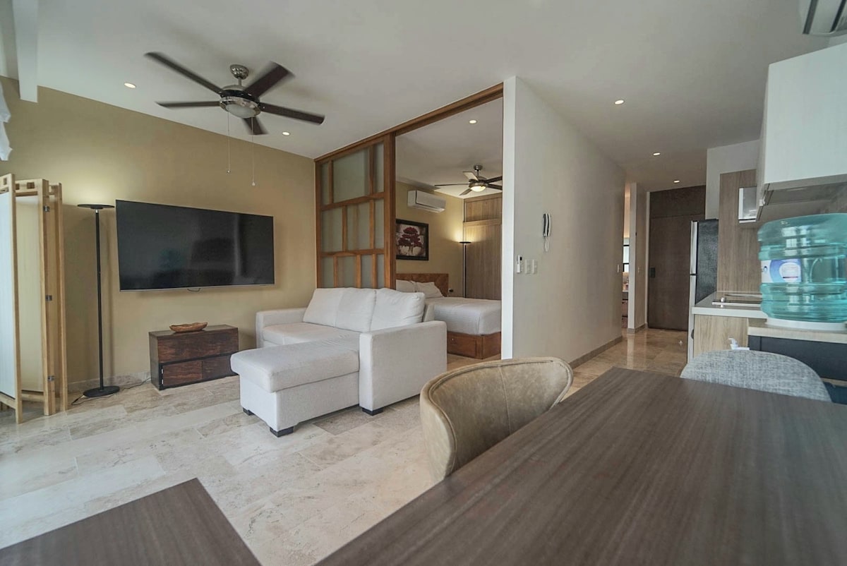 A modern living area displays a light-colored sofa and a large television mounted on the wall. Two ceiling fans provide airflow. A sliding partition separates the bedroom area from the main space, contributing to an open yet defined layout. Natural light enters from various openings.