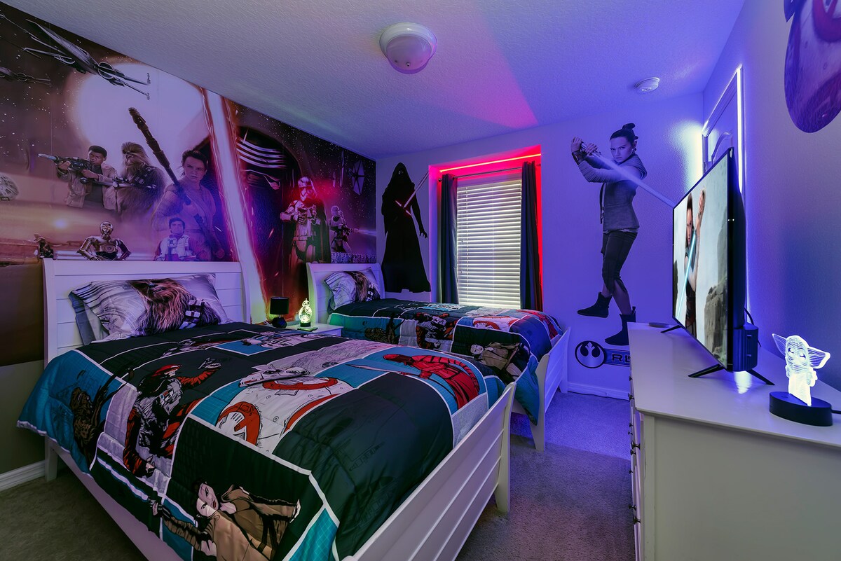 A themed bedroom features two twin beds adorned with Star Wars bedding. Colorful wall art depicts characters from the franchise, while ambient lighting creates a playful atmosphere. A television sits atop a dresser, and a window is dressed with blinds, providing natural light control.