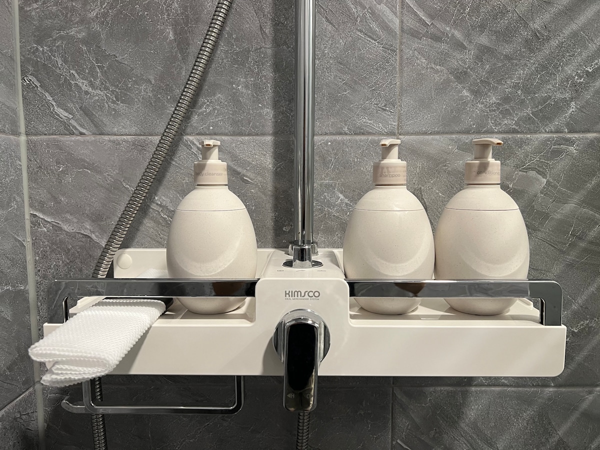 Three pumps of shower amenities are arranged on a sleek, white dispenser mounted on a gray tiled wall. A set of neatly folded disposable washcloths is placed adjacent to the dispenser, providing convenience for guests.