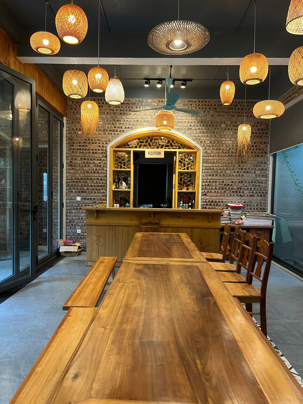 A spacious communal dining area features a long wooden table accompanied by several wooden chairs. Designed with a warm color palette, pendant lights made of woven material hang from the ceiling. A decorative wooden reception area with intricate carvings is positioned in the background.