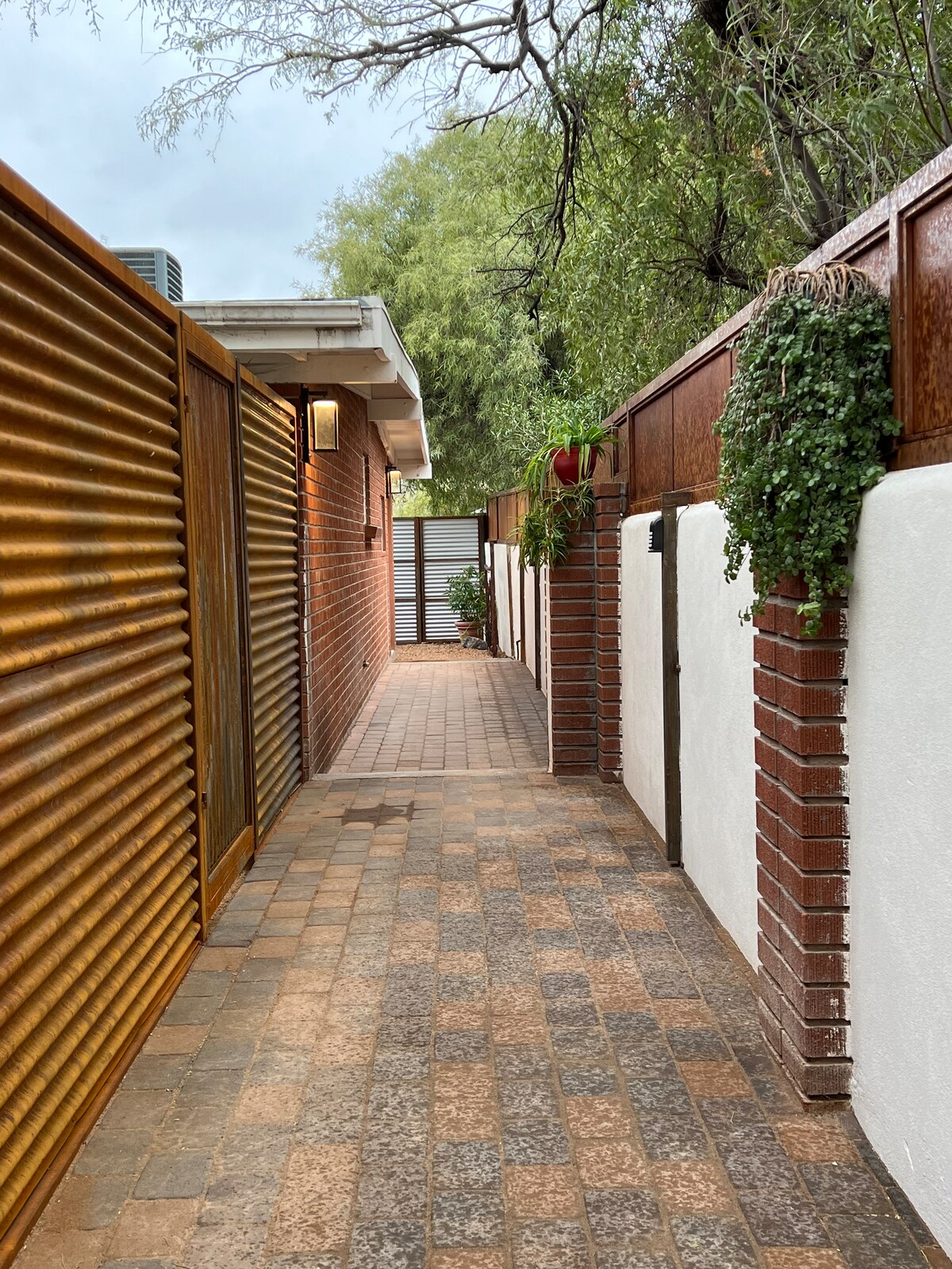 A narrow pathway lined with textured wooden fencing and decorative brick walls guides the way. Lush green plants are seen hanging along the edges, while the peaceful ambiance is enhanced by muted natural light filtering through the spaces.