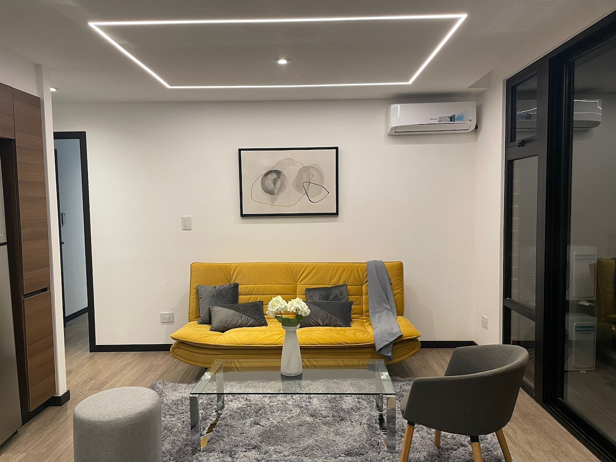 A modern living area features a bright yellow sofa with decorative pillows, positioned in front of a glass coffee table. A soft gray rug covers the floor, while light-colored walls and recessed lighting add to the ambiance. A piece of abstract art is mounted above the sofa.