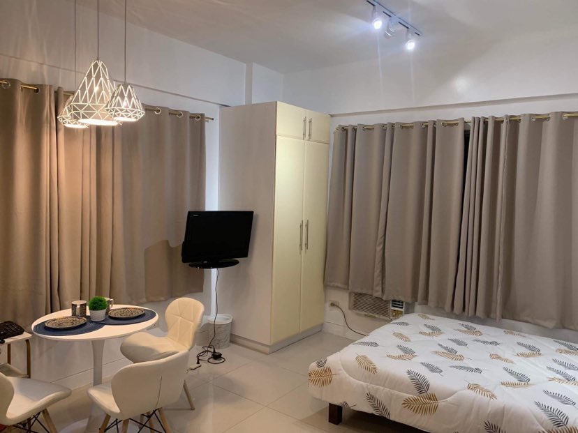 A studio unit is outfitted with a spacious bed adorned with a leaf-patterned blanket. A small dining area features a round table with two chairs, while a wall-mounted television is visible nearby. Soft curtains frame the windows, allowing natural light to filter in.