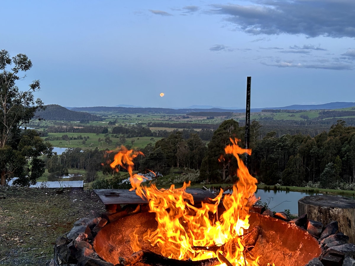 A fire pit is featured in the foreground, flames flickering against a backdrop of expansive rural and mountainous scenery. The landscape includes rolling hills and a distant view of a tranquil water body, with a soft glow from the rising moon illuminating the sky.