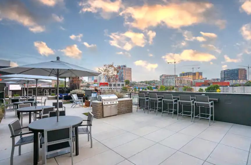 A spacious rooftop terrace is visible, featuring multiple seating areas including tables and barstools. A grill is positioned for outdoor cooking, with a backdrop of the Nashville skyline against a partly cloudy sky.