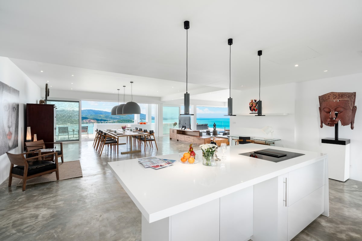 The open-concept living and dining area features a modern kitchen island with sleek surfaces. Large windows provide ample natural light and showcase views of the ocean. Stylish wooden chairs surround the dining table, while minimalist decor enhances the spacious atmosphere.