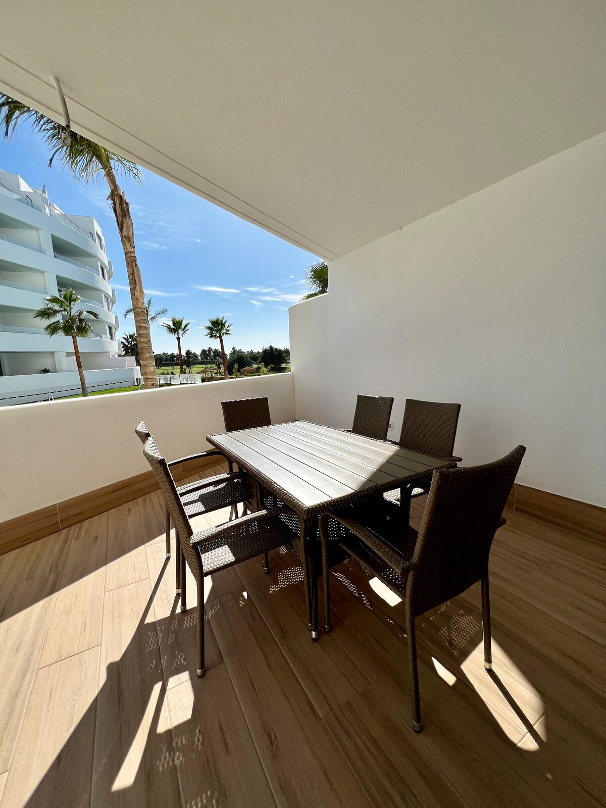 A terrace is presented with a large dining table surrounded by six woven chairs. Sunlight illuminates the space, highlighting the clean lines of the white walls and the warm tones of the wooden flooring. Palm trees are visible in the background, fostering a tranquil environment.