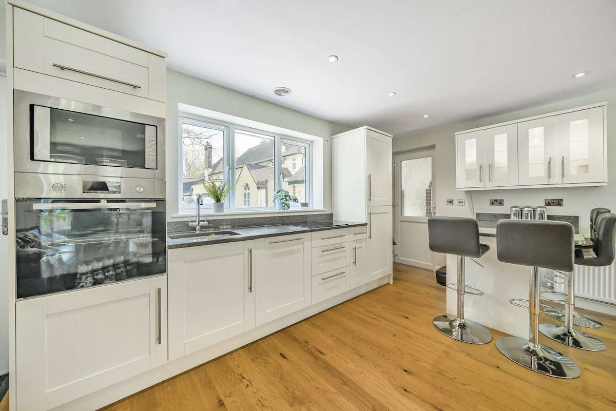 A modern kitchen space features white cabinetry and granite countertops, providing ample storage and preparation area. Stainless steel appliances are integrated seamlessly. A breakfast bar with high stools offers additional seating, while natural light floods the room through large windows.