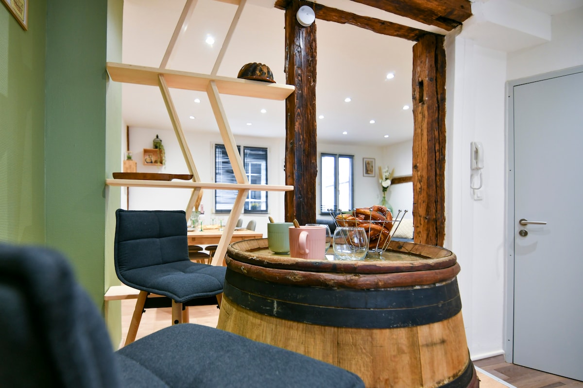 A wooden barrel serves as a unique table, complemented by two modern chairs in a cozy, inviting setting. Exposed wooden beams highlight the architectural character of the space, while soft lighting enhances the overall ambiance, creating a welcoming atmosphere for relaxation.