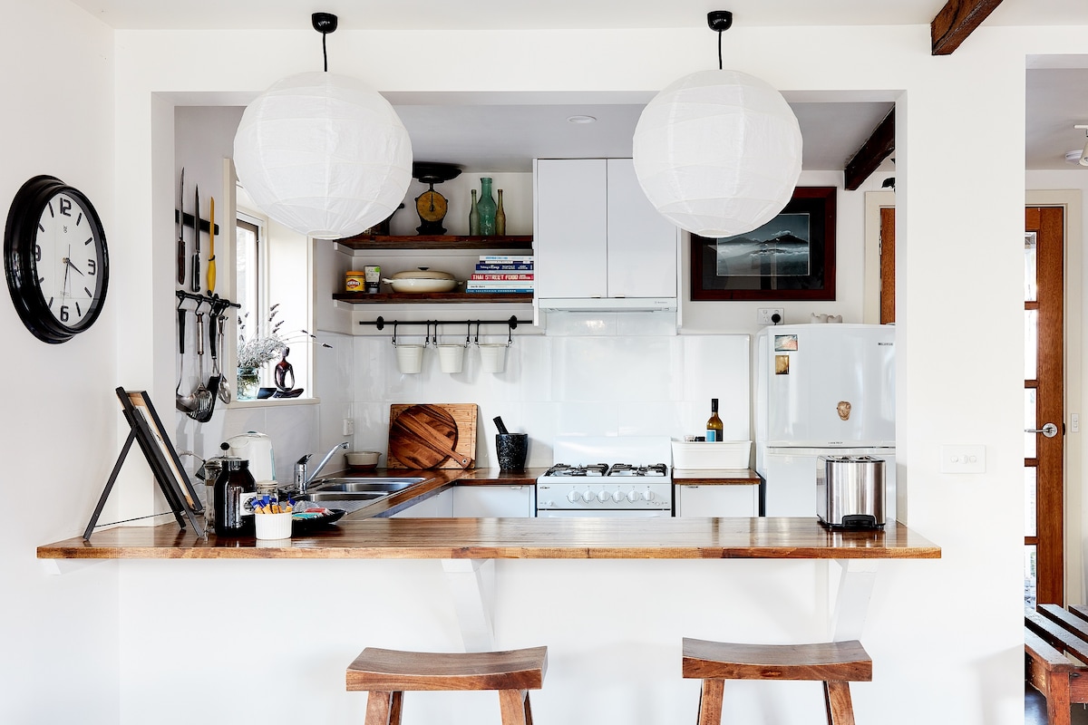 A functional kitchen space is highlighted by two pendant lights hanging above a wooden breakfast bar with two stools. The area features a gas stove, sink, and white appliances, with utensils displayed on the shelves above, creating a well-organized and welcoming cooking environment.