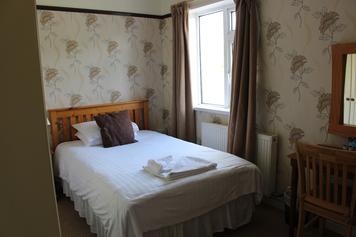 A cozy sleeping area features a spacious bed adorned with white linens and a brown accent pillow. Natural light enters through a large window dressed with beige curtains. A wooden desk and mirror are positioned nearby against a floral-patterned wallpaper.
