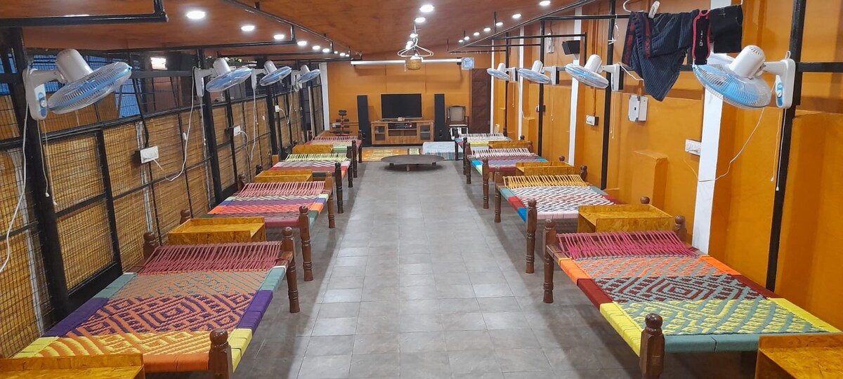 A spacious room features multiple thread-woven cots arranged neatly along the sides, with vibrant patterns. A television can be seen in the center, flanked by ceiling fans and wall-mounted lights. The warm-toned walls contribute to a cozy, relaxed environment.