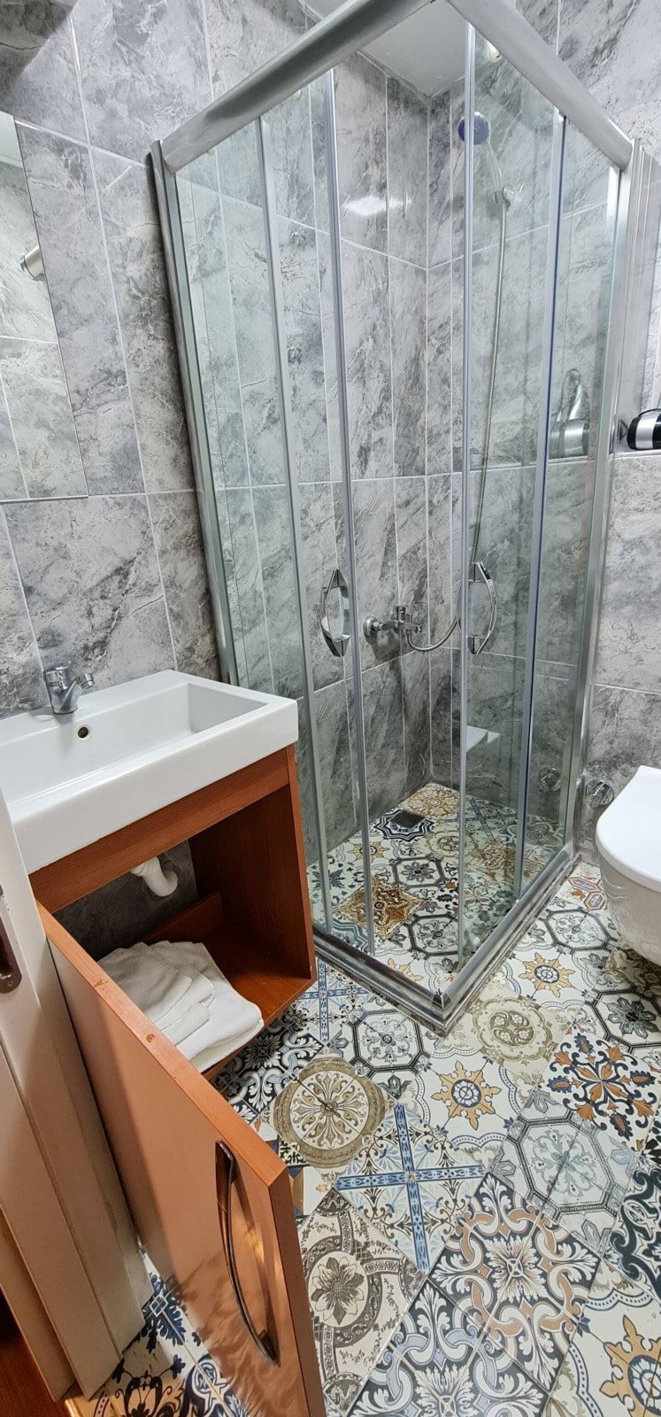 A modern bathroom features a glass-enclosed shower with dual shower heads. A sleek sink is positioned on a wooden cabinet, complemented by colorful patterned tiles on the floor. Basic toiletries are neatly stored in the cabinet.