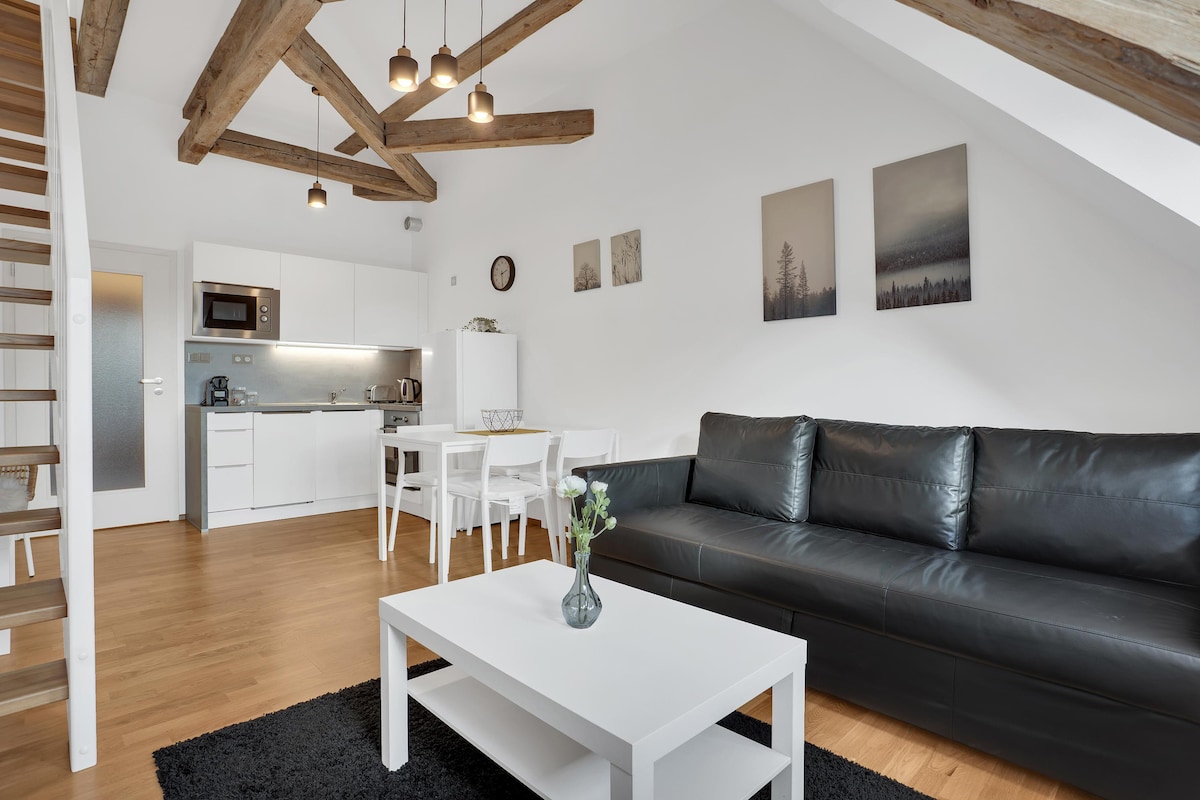 A modern living area features wooden beams overhead and large windows allowing natural light. A spacious black couch faces a white coffee table, with a vase of flowers displayed. A dining table with four chairs is positioned beside a fully equipped kitchen.