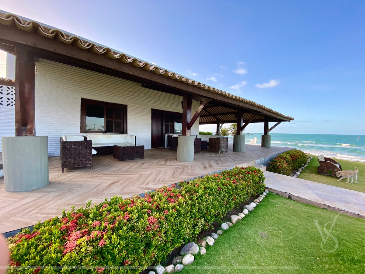 An outdoor patio features a spacious wooden deck with ample seating provided by woven chairs. Lush, colorful landscaping lines the edge, while the tranquil sea can be seen in the distance, contributing to a serene atmosphere.
