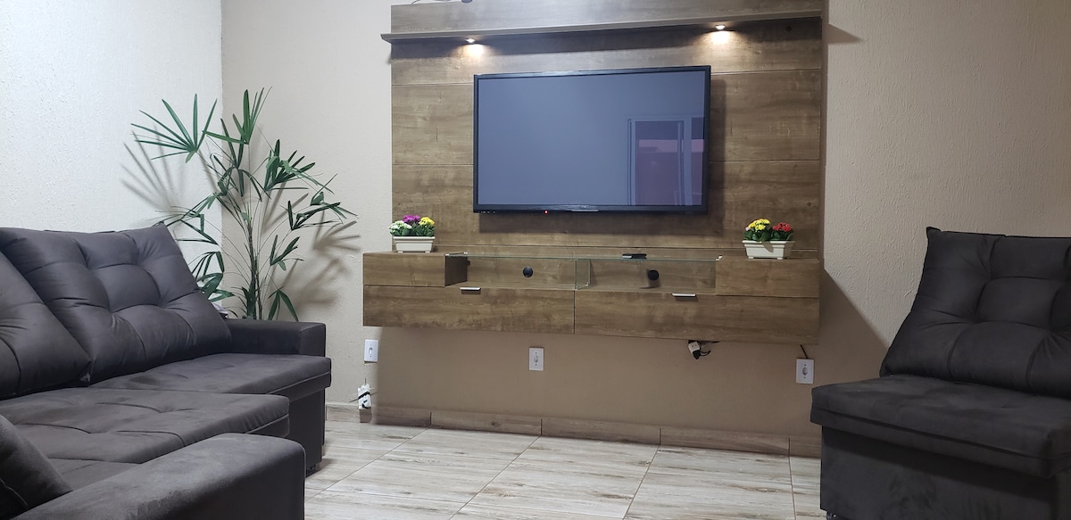A spacious living room is presented, featuring two plush sofas arranged around a central area. A flat-screen television is mounted on a wooden panel, while decorative plants and flower pots add a touch of greenery. Soft lighting highlights the warm tones of the furnishings.