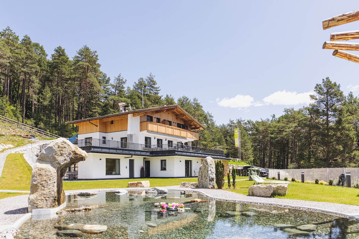 A modern two-story building is set among trees, featuring wooden accents on the upper level. A tranquil pond is visible in the foreground, surrounded by smooth stones and adorned with flowering plants. A clear sky enhances the natural light over the inviting landscape.