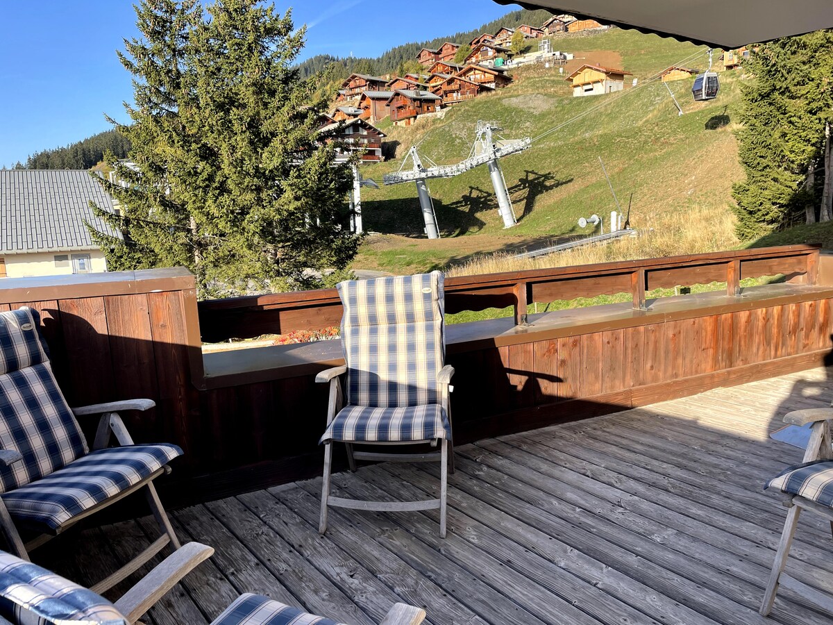 A wooden terrace is presented, featuring four comfortably spaced lounge chairs with checkered fabric. The surrounding landscape includes greenery and a ski lift, with quaint wooden chalets situated on the hillside in the background.