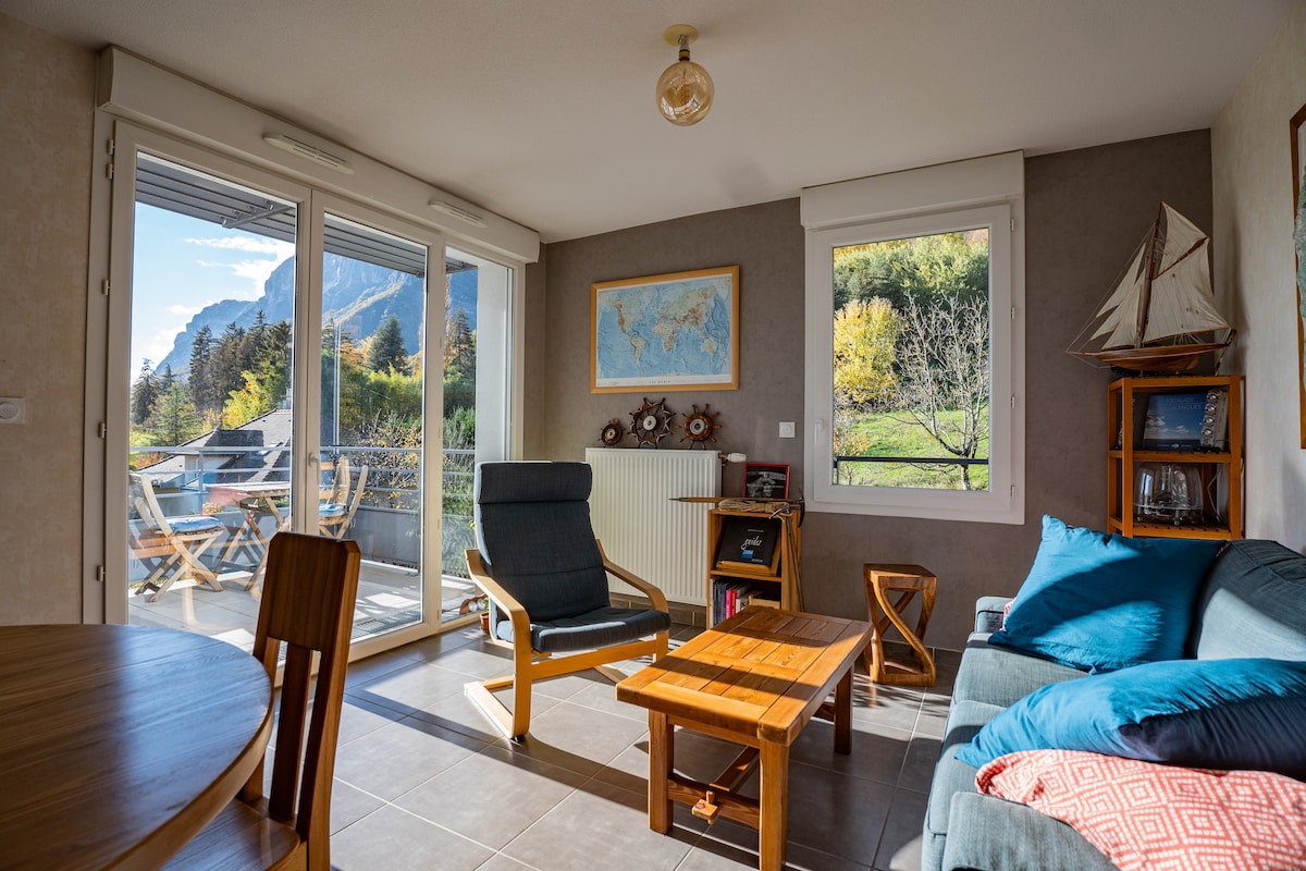 A bright living area features large glass doors that open to a terrace with a mountainous view. A comfortable blue sofa and a wooden coffee table create a cozy space, complemented by a simple dining table and a stylish chair. Natural light fills the room.