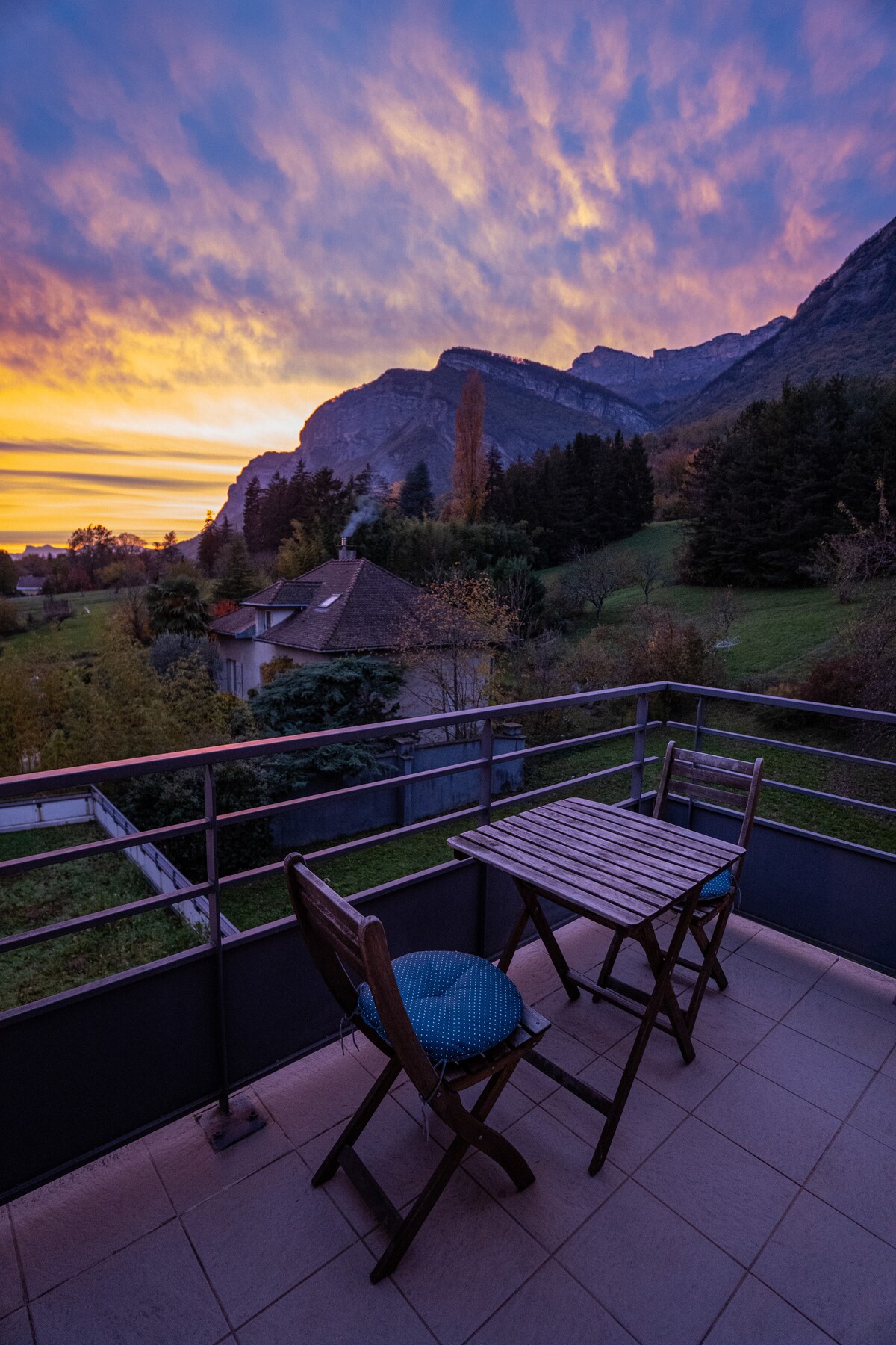 A terrace is shown with a wooden table and two chairs, offering a view of the surrounding mountains at sunset. The sky is filled with vibrant colors, and lush green hills are visible in the background, creating a serene outdoor space.