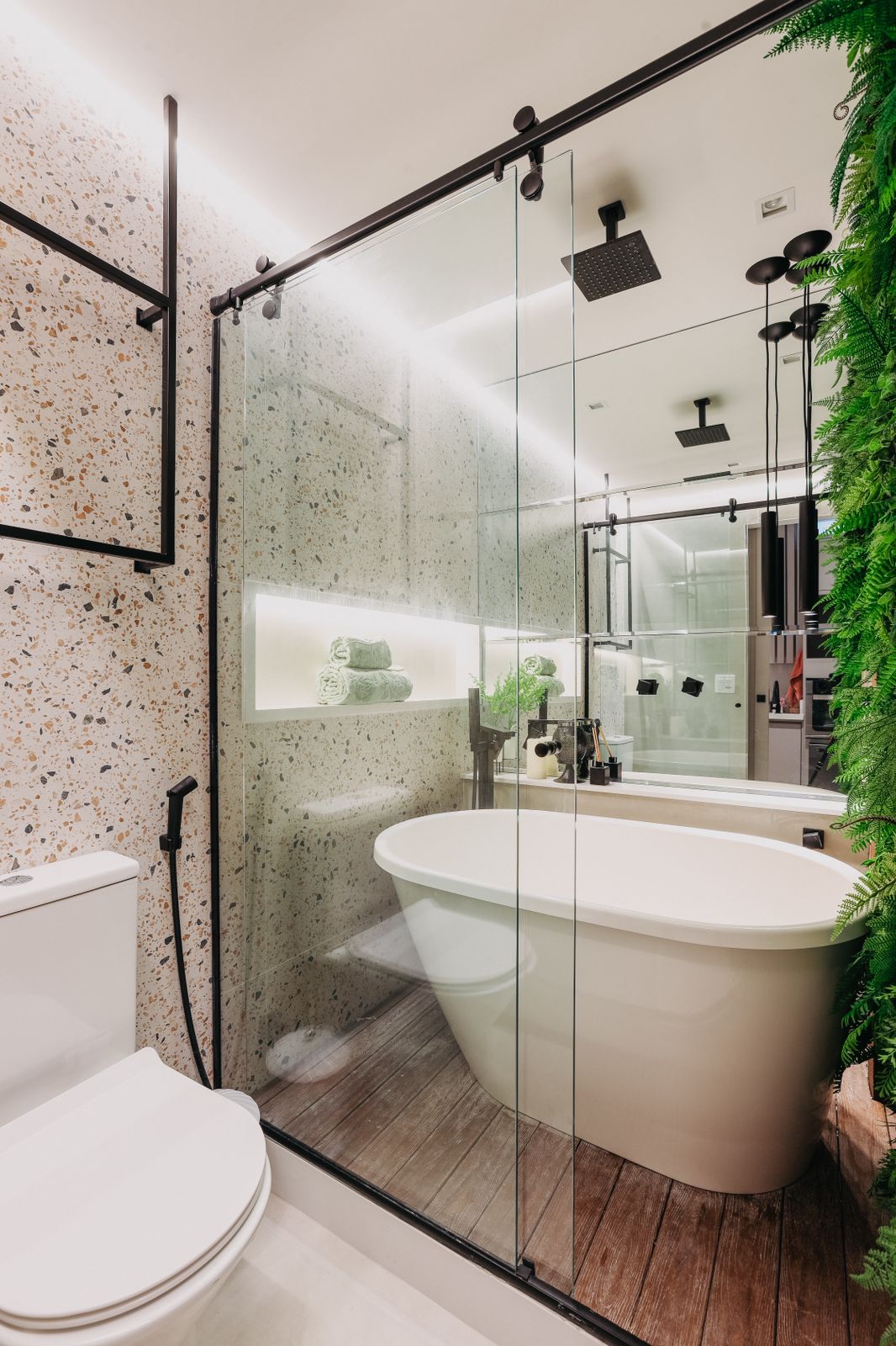 The bathroom features a spacious soaking tub enclosed by glass, complemented by a minimalist design. Textured walls add warmth, and a well-placed shelf holds neatly arranged towels. Modern lighting fixtures enhance the ambiance, while greenery provides a refreshing touch.