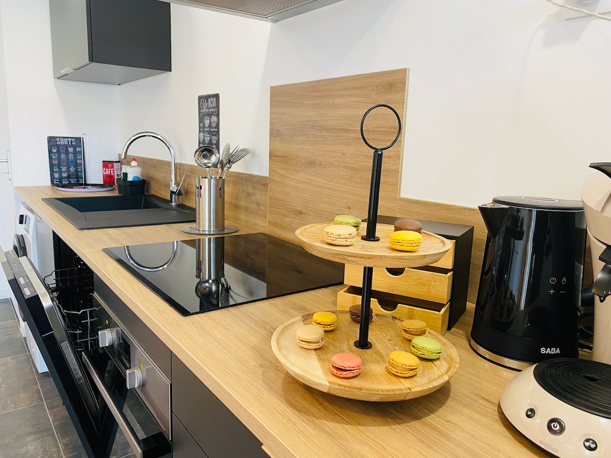 A modern kitchen showcases sleek black cabinetry and a wooden countertop. An electric kettle and coffee machine sit alongside a stylish two-tiered stand adorned with colorful macarons. Cooking tools and an induction cooktop are also visible, creating a functional and inviting space.