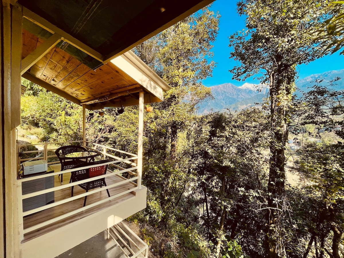 A shaded terrace features two comfortable chairs overlooking a lush forest. The scenic view includes vibrant green trees and distant mountains under a clear blue sky, creating a serene outdoor space for relaxation.