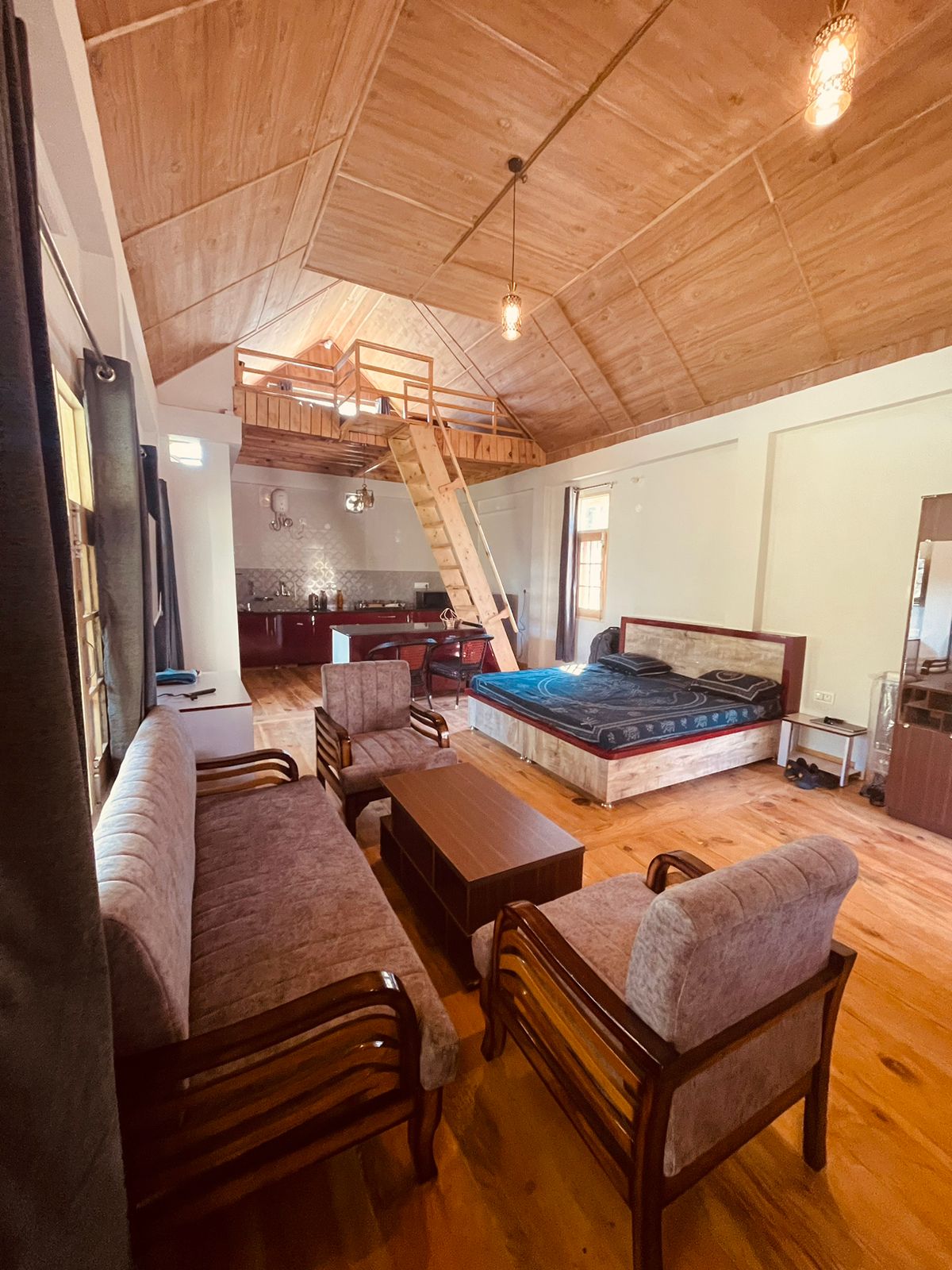 An airy interior showcases a spacious seating area with two sofas and two armchairs, complemented by a wooden coffee table. A large bed is positioned in the corner, and a staircase leads to a cozy attic. Natural light filters through the windows, illuminating the wooden ceiling.