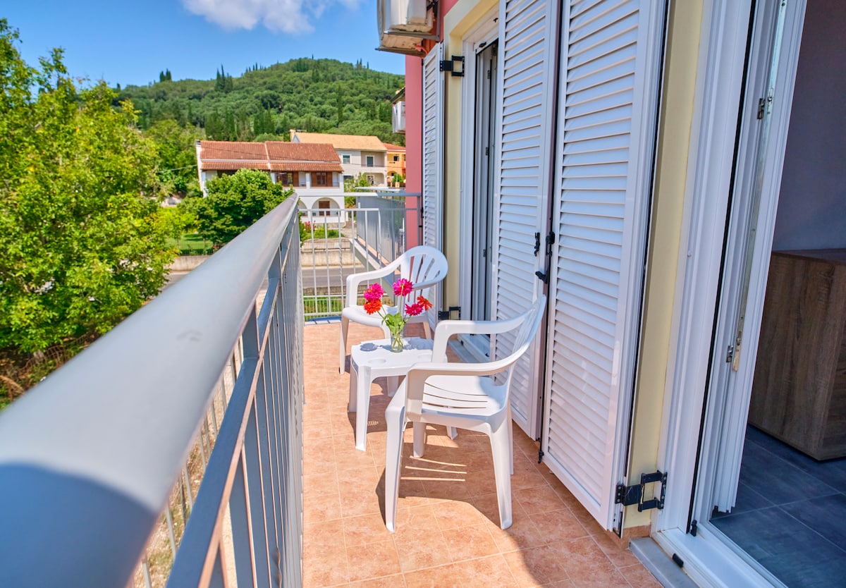 A balcony features two white plastic chairs and a small table adorned with colorful flowers. The surrounding landscape includes lush greenery and neighboring buildings, with sunlight illuminating the space. Large glass doors provide access to the indoors, enhancing the open atmosphere.