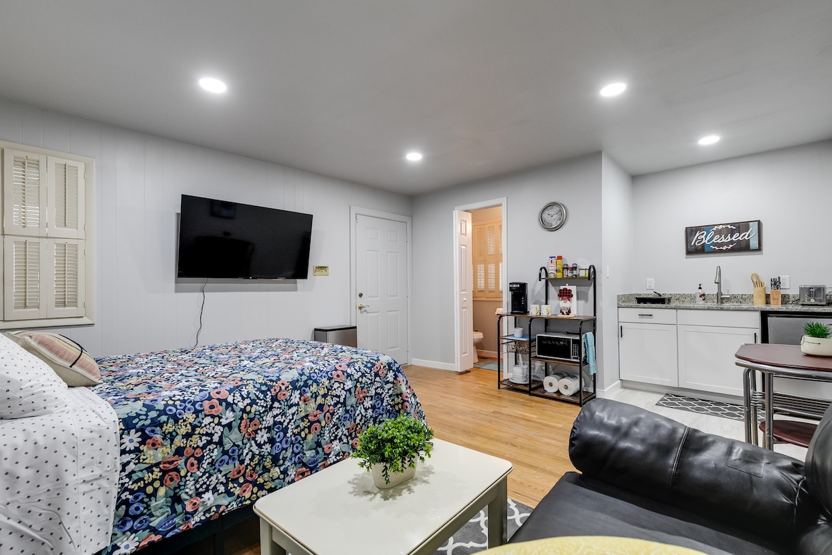 A cozy studio space is equipped with a spacious bed adorned with floral bedding, complemented by a large flat-screen TV mounted on the wall. A kitchenette with modern appliances is visible, alongside a small dining table and a comfortable seating area.