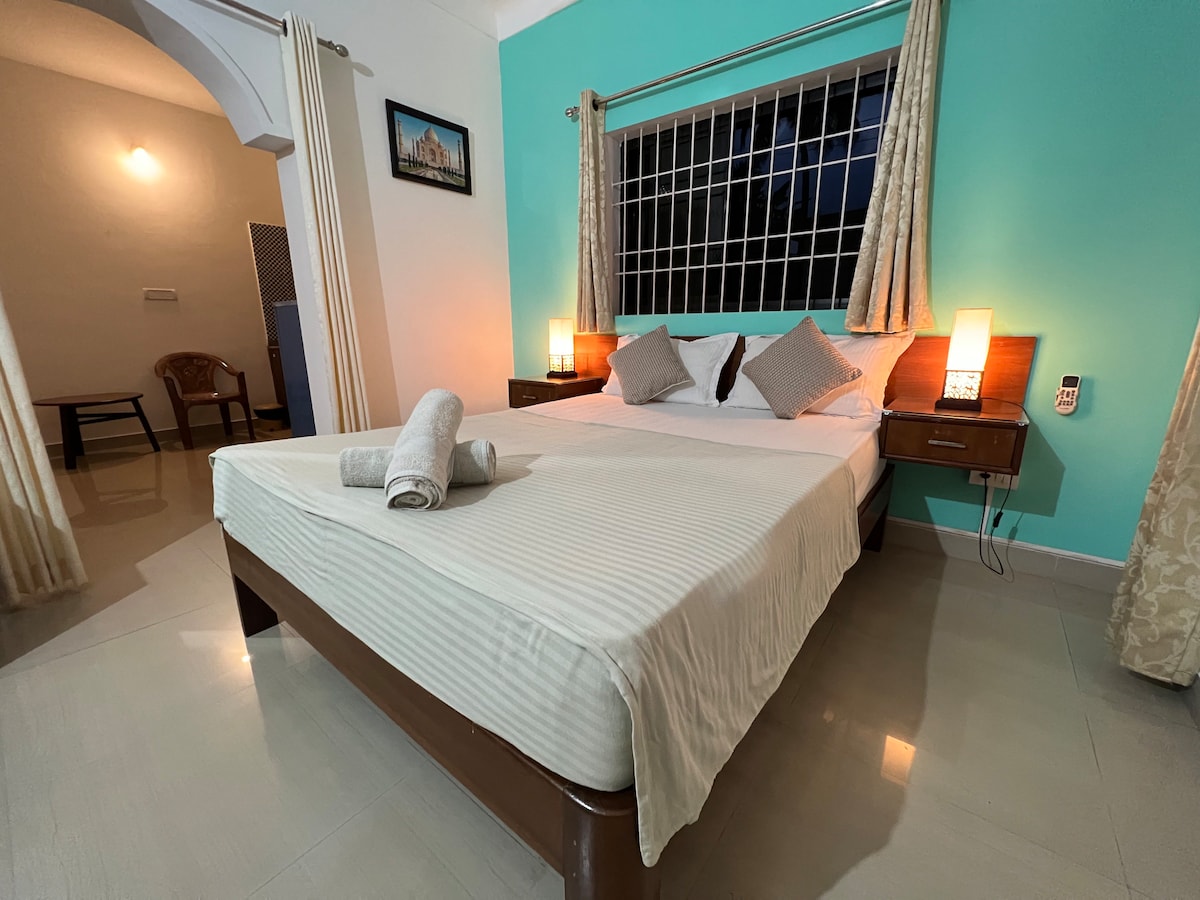 A well-lit sleeping area features a spacious bed with light-colored linens. Two decorative pillows add a touch of style. Soft lamps illuminate the room, contrasting with the turquoise wall. A large window provides natural light, framed by light curtains.