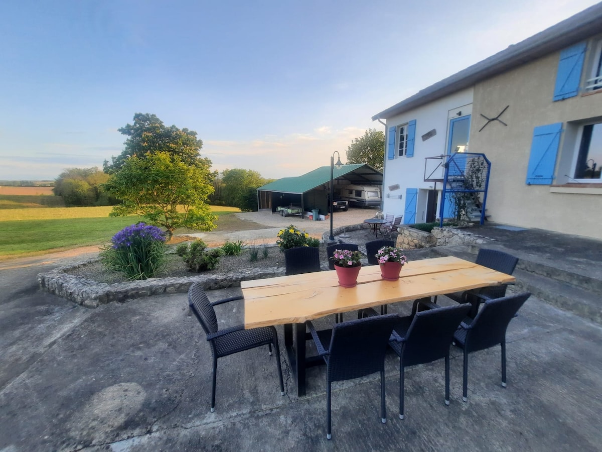 An outdoor dining area features a long wooden table surrounded by six black chairs. Potted flowers add color to the table. A grassy expanse and trees are visible in the background, with a covered parking area further back, complementing the peaceful rural setting.