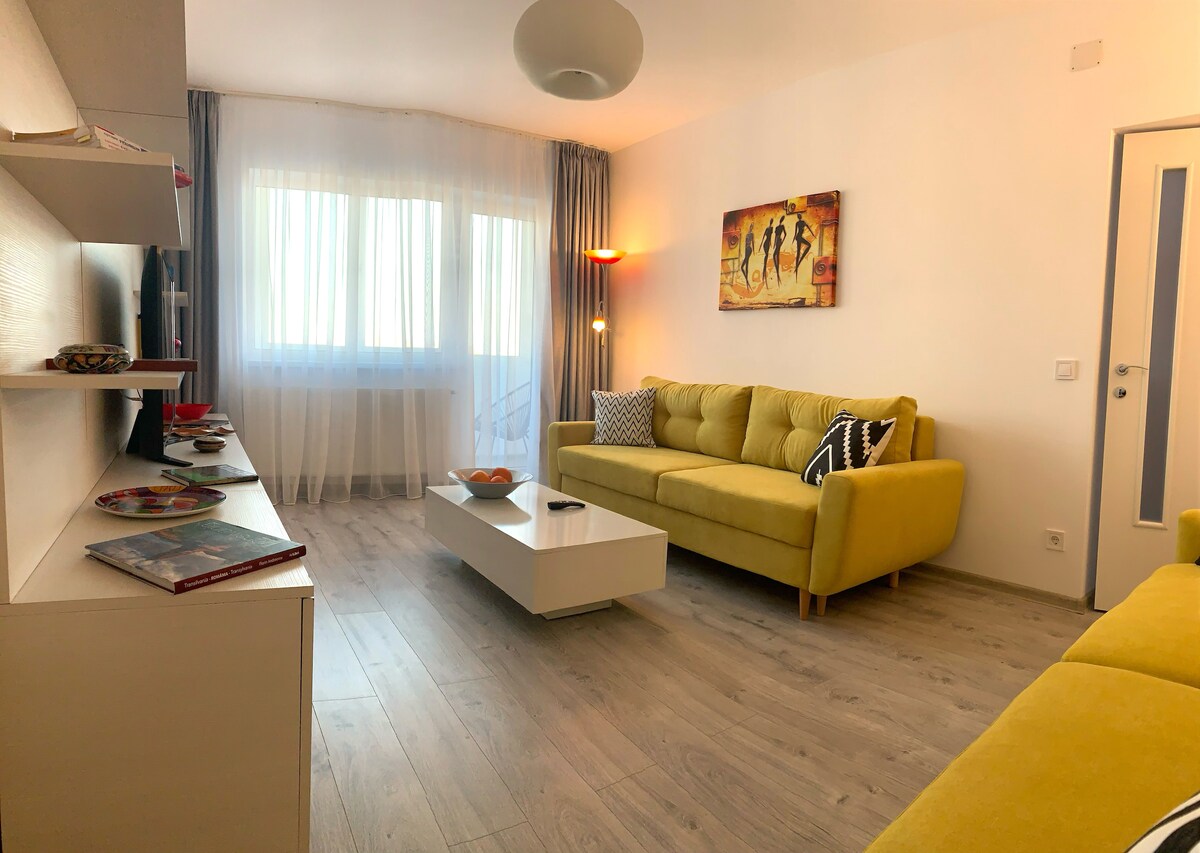 A modern living area features a bright yellow sofa and a white coffee table. Natural light enters through large windows covered by sheer curtains. A piece of abstract art adorns the wall, complemented by a stylish lamp that provides additional lighting.