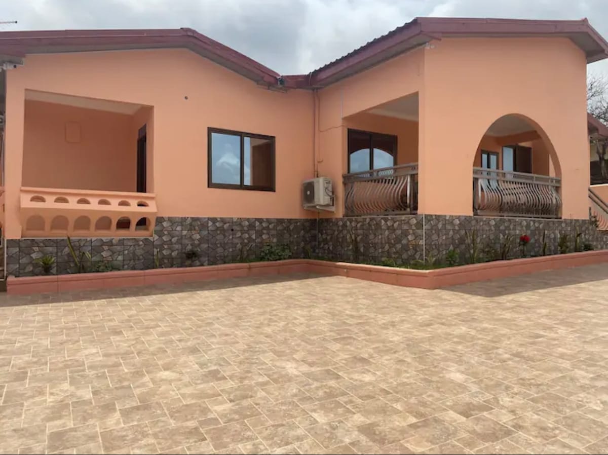 A single-story building with a peach exterior is surrounded by a well-maintained stone wall. Large windows provide natural light, while a decorative arch leads to a covered balcony area. An outdoor air conditioning unit is mounted on the side, and the driveway is paved with light-colored tiles.