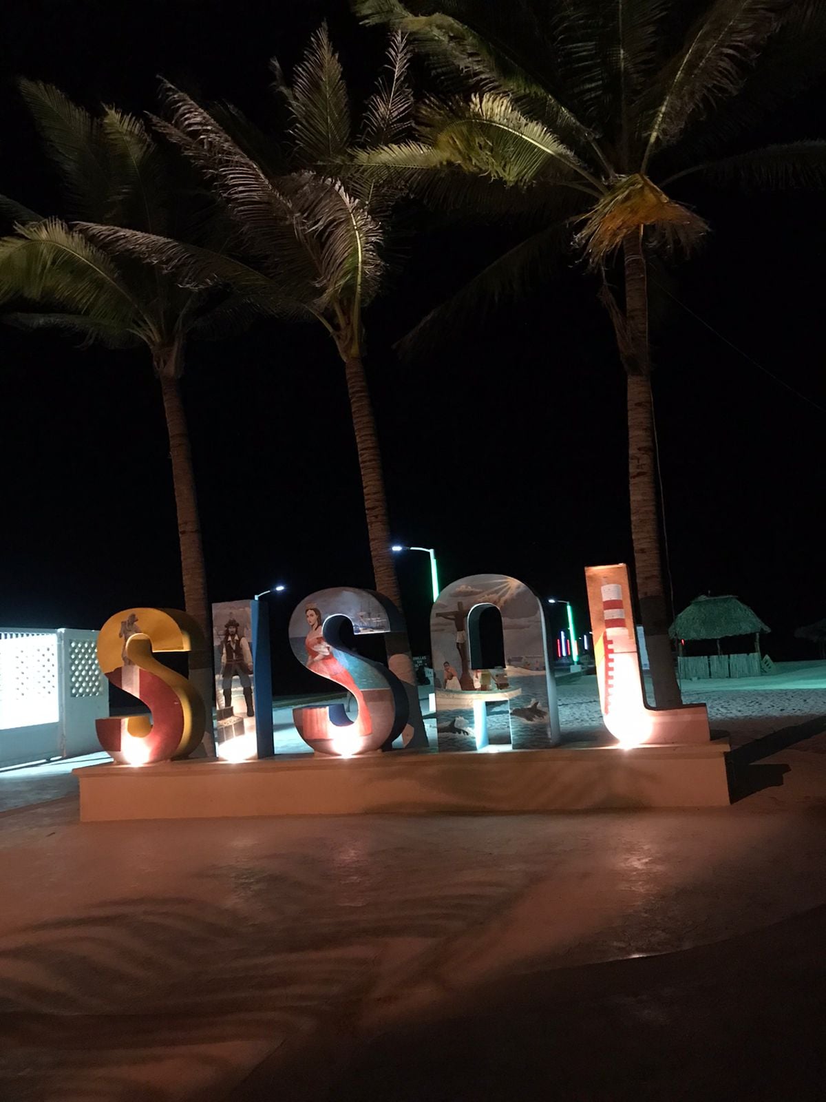 Colorful letters spelling 'Sisal' are illuminated at night, surrounded by palm trees swaying softly in the breeze. A serene beachfront setting is visible in the background, adding to the peaceful ambiance of the area.
