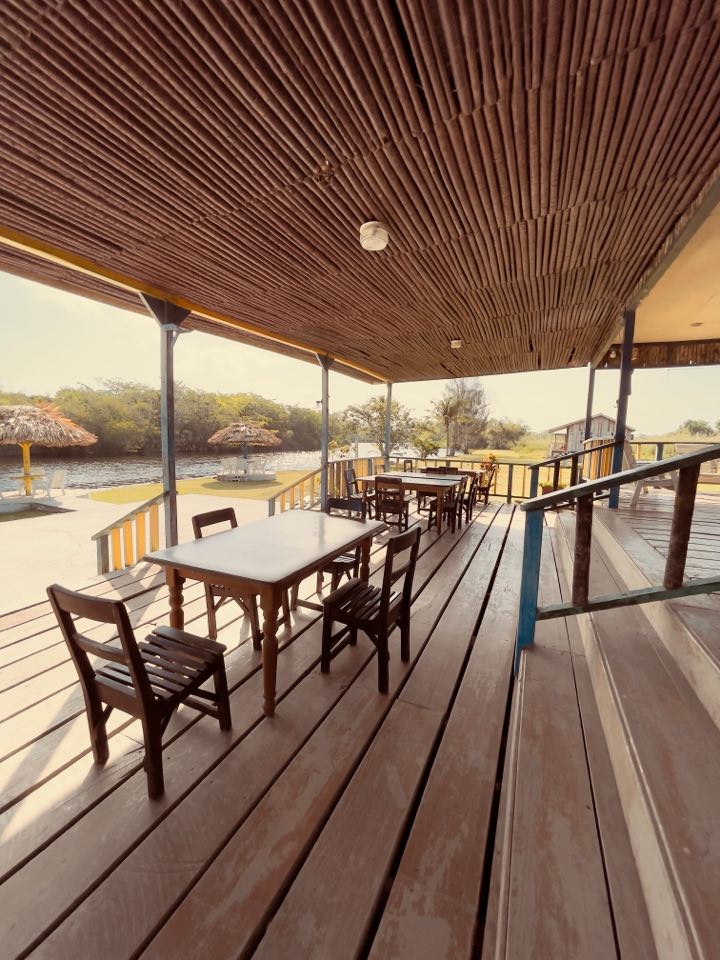 An outdoor dining area features several wooden tables and chairs under a thatched roof. The floor consists of broad wooden planks, and sunlight fills the space, highlighting the open view of the riverside and surrounding greenery.