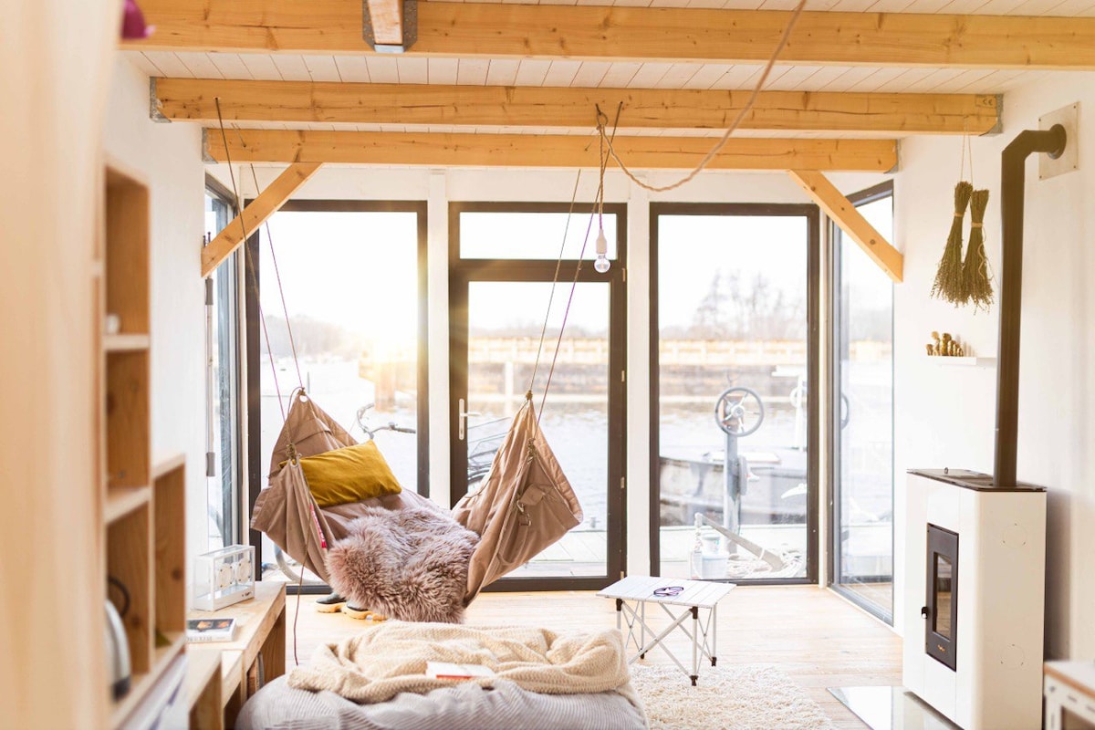 A cozy living space features large glass doors allowing natural light to fill the room. A softly textured area is complemented by a hammock suspended from wooden beams. A white stove stands next to a simple coffee table, enhancing the comfortable ambiance.