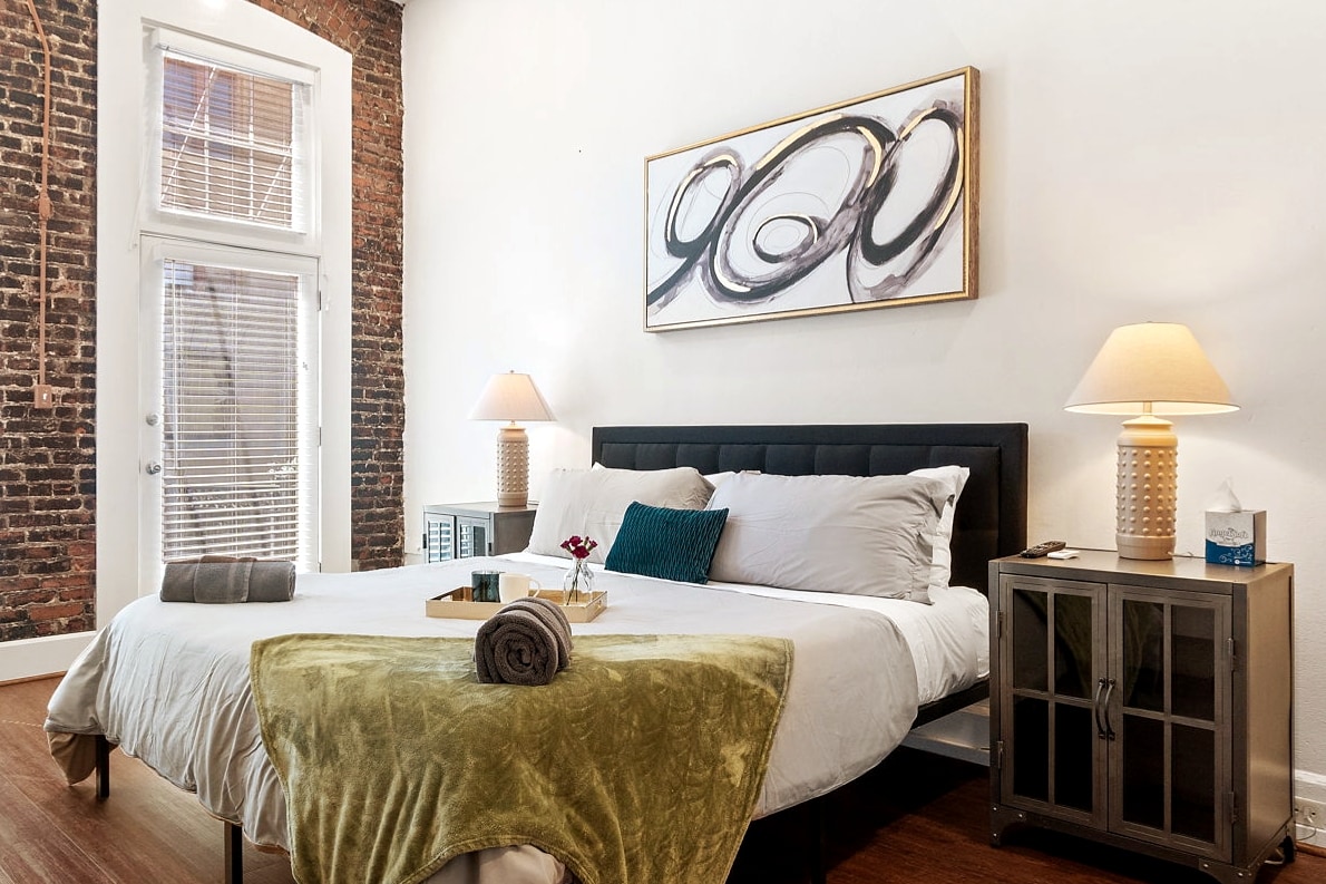 A spacious bedroom features exposed brick walls and large windows allowing natural light. The king-sized bed is dressed in soft linens with decorative pillows. Nightstands with warm lamps flank each side, while a small vase holds flowers on the bed, enhancing the inviting atmosphere.