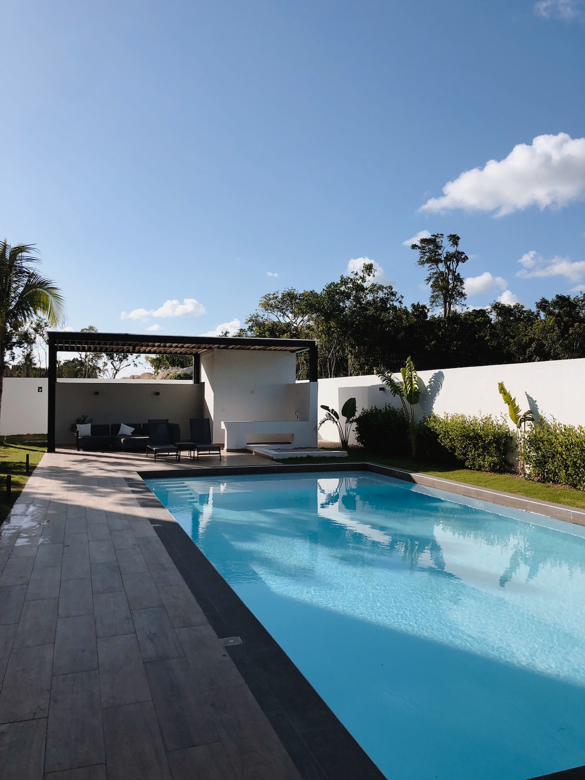 A wide swimming pool is presented, surrounded by a wooden deck and well-tended greenery. A shaded seating area with modern furniture is visible nearby, complemented by a clean, white wall that integrates with the landscape. Bright skies and sunlight add to the serene environment.