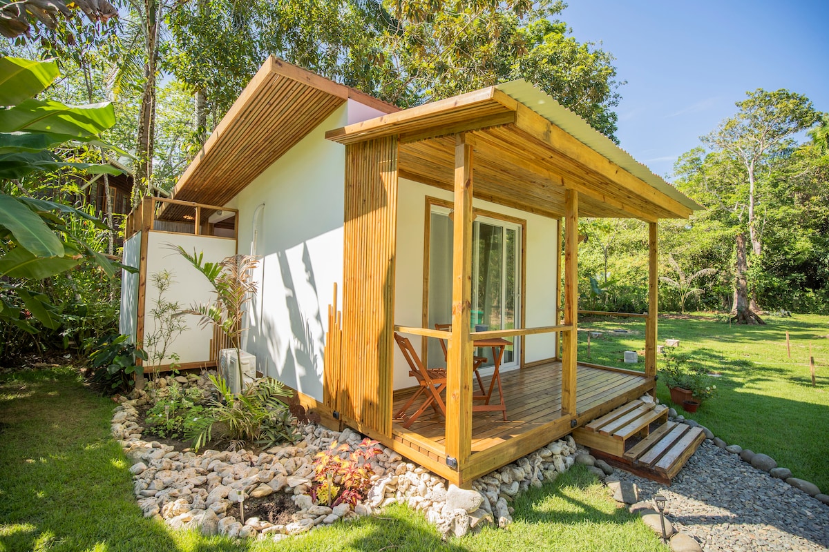 A charming cabin is depicted, featuring a wooden porch with two outdoor chairs and a table. Lush greenery surrounds the structure, and a stone pathway leads to the entrance. Large windows allow natural light to illuminate the space.