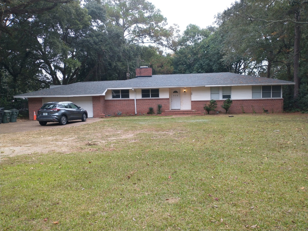 A single-story brick home is set on a grassy lot, framed by trees. A vehicle is parked in front, and a path leads to the welcoming entrance. The exterior features a mix of brick and light siding, with a centered front door and two small windows on either side.