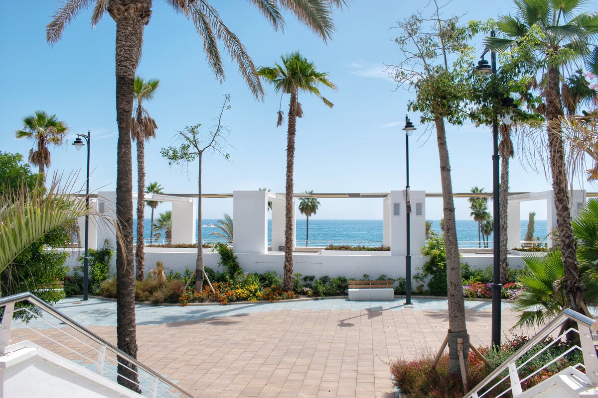 A sunlit coastal view showcases a vibrant scene framed by palm trees and flowering plants. In the distance, the sea glimmers under bright blue skies, providing a serene backdrop to the inviting outdoor space.