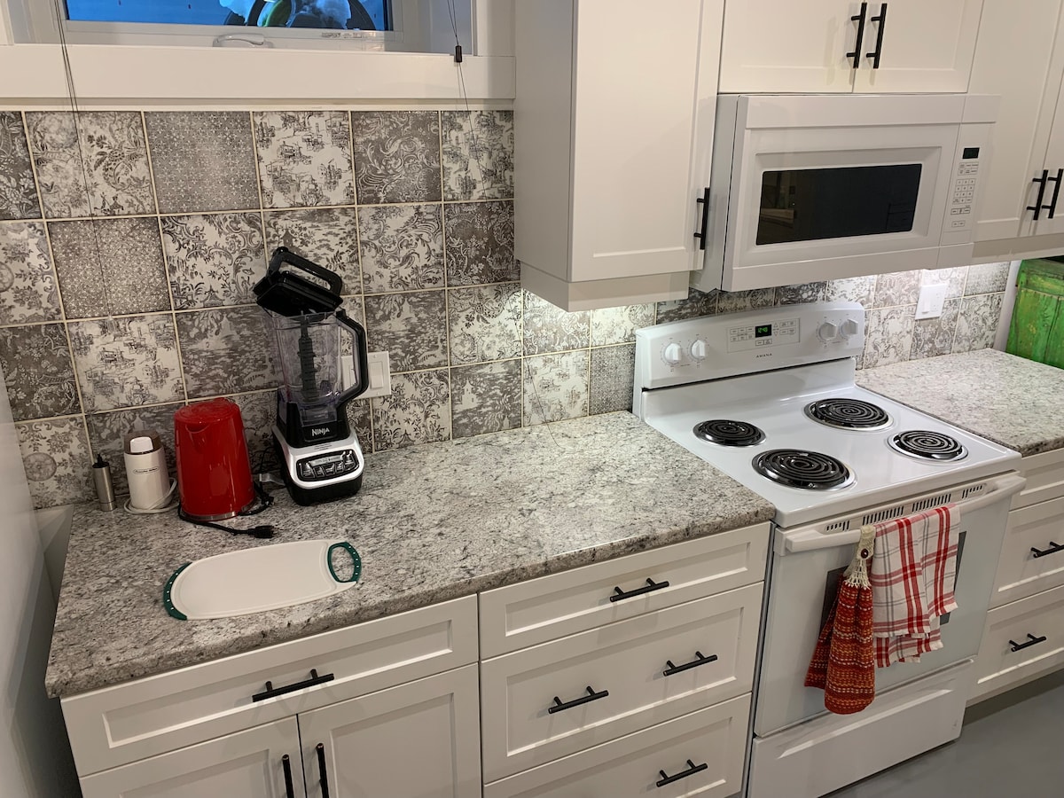 A modern kitchen features a granite countertop with a blender, coffee maker, and microwave positioned above a stove. Decorative tile backsplash provides texture, while white cabinetry offers ample storage. Soft lighting enhances the space, creating an accessible environment for meal preparation.