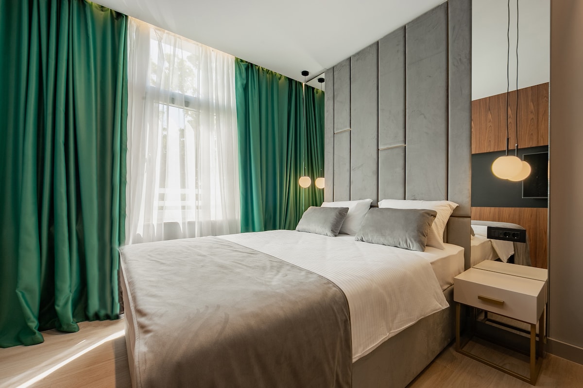 A cozy bedroom features a spacious bed with neutral bedding, complemented by soft gray throw pillows. Rich green curtains frame the large window, allowing natural light to filter through. Modern bedside tables with stylish lamps add an elegant touch to the tranquil setting.