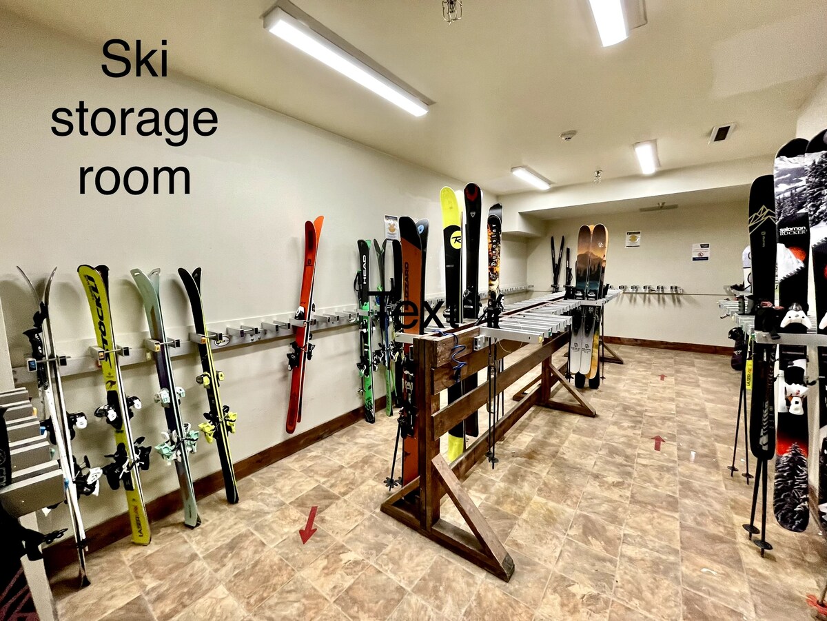 The ski storage room features wooden racks positioned for efficient organization of skiing equipment. Multiple pairs of skis are arranged along the walls, providing ample space for storage. The flooring is tiled and lights illuminate the area, ensuring visibility.