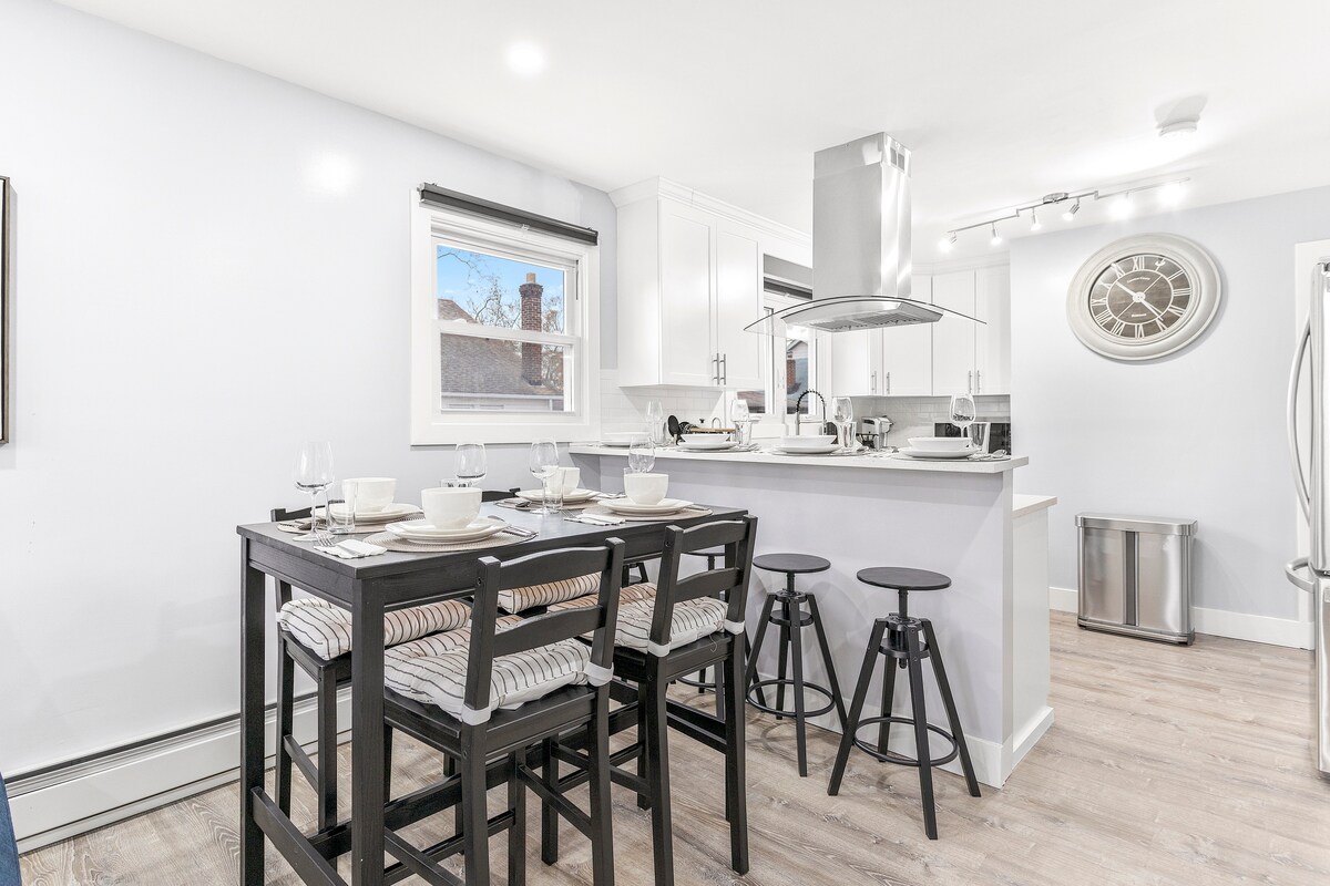 An open-concept kitchen and dining area features a modern, well-equipped kitchen with stainless steel appliances. A black dining set with four place settings and stylish bar stools creates a welcoming atmosphere. Natural light floods the space through a window, highlighting the sleek design.