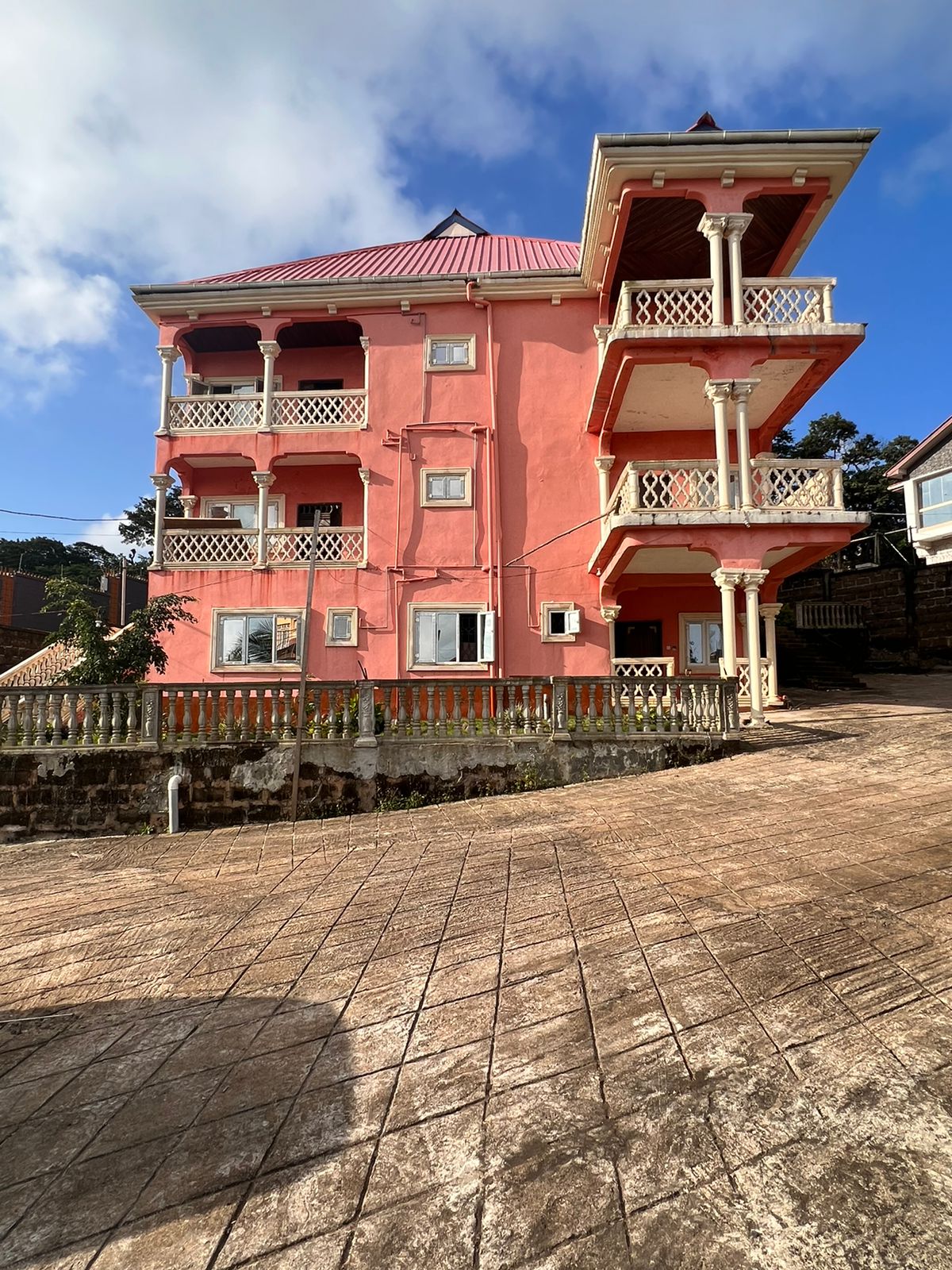 The exterior of a vibrant pink house is displayed, featuring four levels, with decorative balconies and white railings. The structure is situated on an incline, surrounded by greenery. A clear blue sky complements the colorful design, highlighting the distinct architectural style.